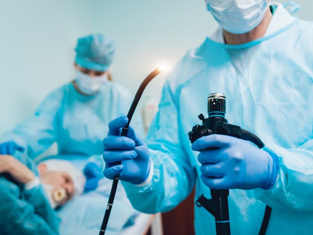 Profissional segurando o endoscópio pronto para iniciar o exame, com paciente sedado ao fundo. O resultado da endoscopia sai na hora para avaliações visuais.