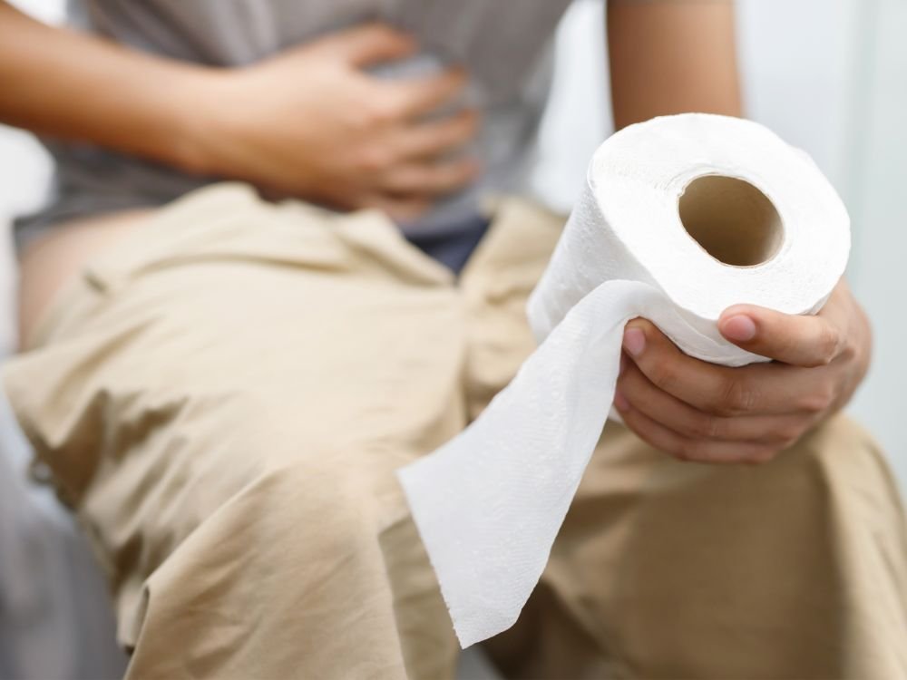 Pessoa sentada no vaso sanitário com dor abdominal e papel higiênico na mão, possivelmente com fezes finas.