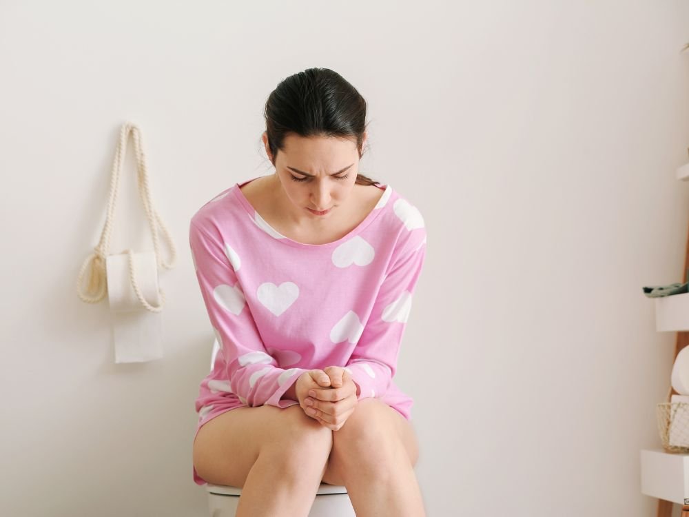 Mulher sentada em uma privada, com uma expressão de desconforto ou dor de estômago, olhando para baixo. A imagem simboliza as possíveis causas médicas para fezes verdes, como infecções ou doenças gastrointestinais.