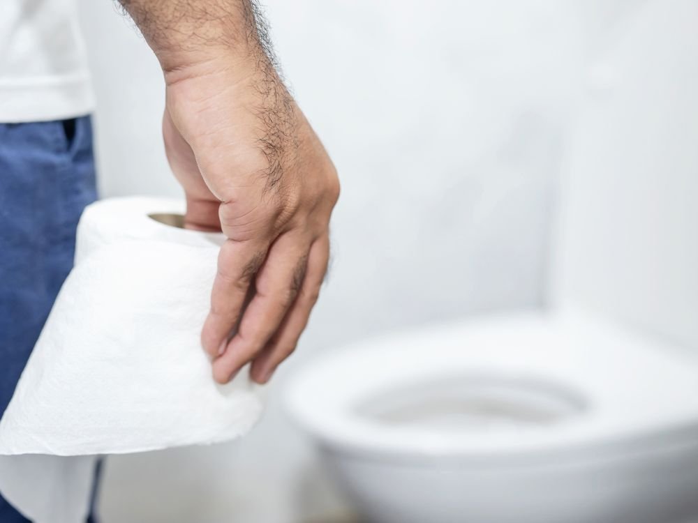 Homem de pé em um banheiro, segurando um rolo de papel higiênico, com o vaso sanitário desfocado ao fundo. A imagem ilustra uma situação comum de preocupação com a saúde intestinal e a aparência das fezes verdes.