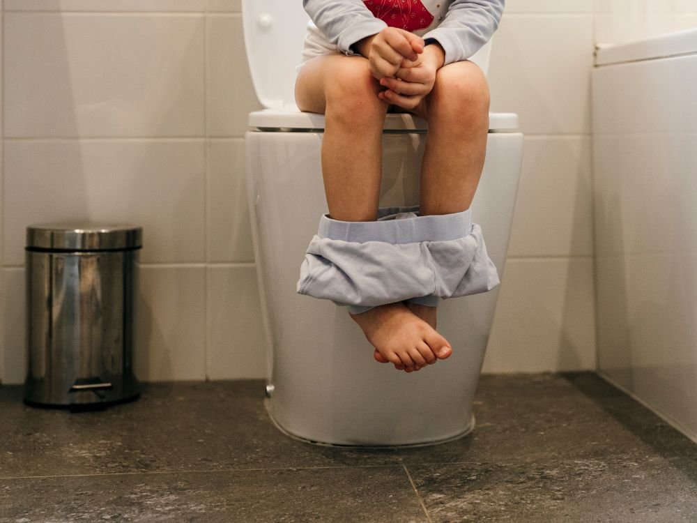 Criança pequena sentada em um vaso sanitário, com as calças abaixadas até os tornozelos. A imagem representa o tema de fezes verdes em crianças e como o trânsito intestinal acelerado pode ser uma causa.