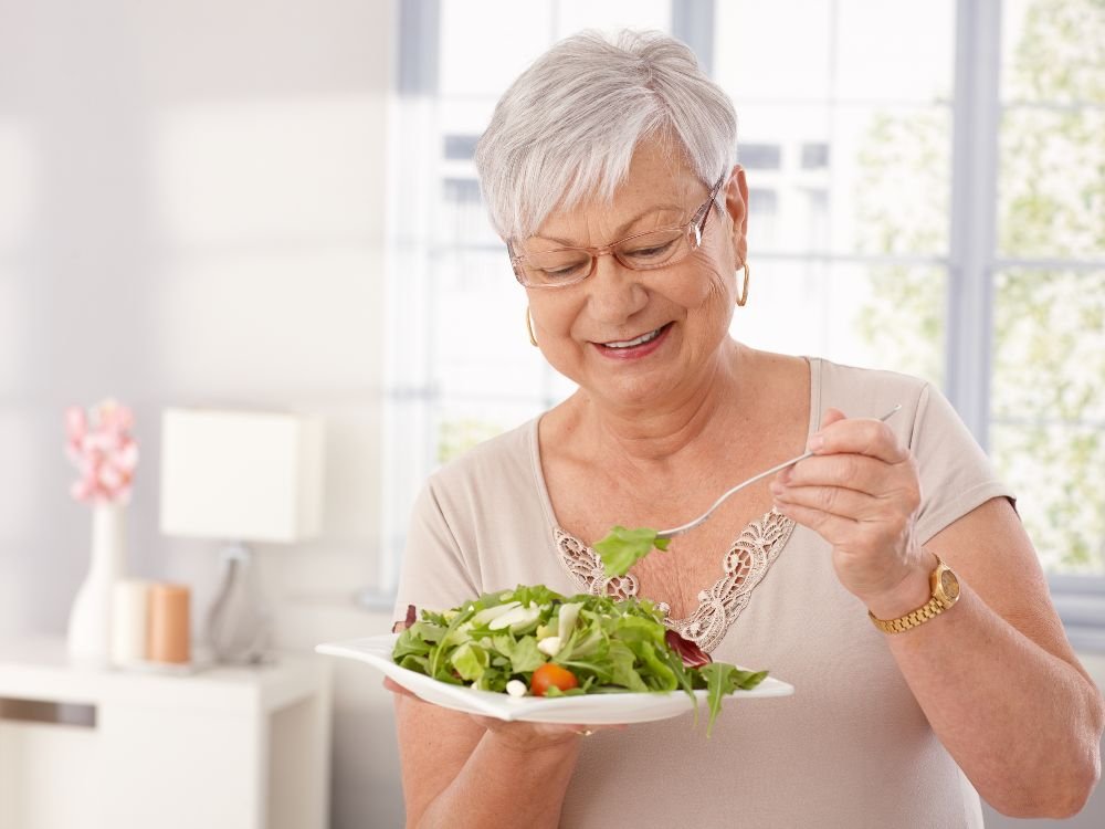 Mulher idosa sorrindo, comendo uma salada de folhas verdes. A imagem destaca uma causa comum e inofensiva para a cor das fezes verdes: o consumo de alimentos ricos em clorofila ou com corantes artificiais.