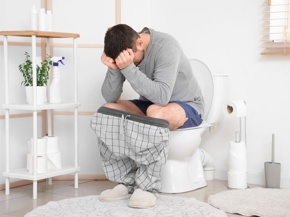 Homem sentado no vaso sanitário com desconforto abdominal, indicando dificuldade para evacuar e presença de fezes em formato de fita.