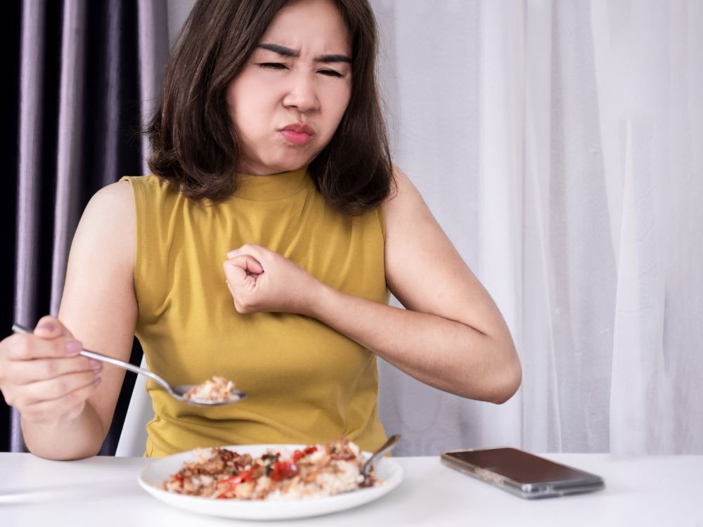 Mulher sentindo dificuldade para engolir alimentos durante refeição, com sensação de desconforto na região do peito.