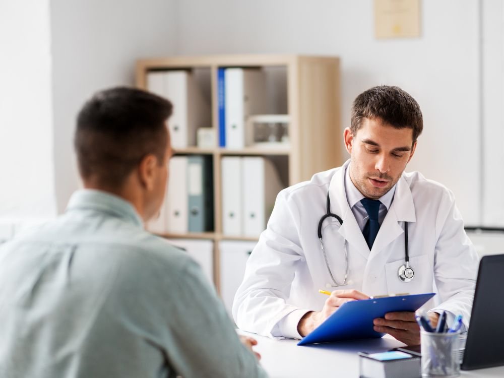 Médico realiza atendimento clínico presencial enquanto conversa com paciente em consultório, avaliando informações e preenchendo prontuário médico.