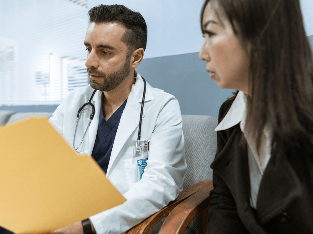 Médico de jaleco branco analisa documentos durante uma consulta com uma mulher, em ambiente clínico, para discutir sintomas de saúde.