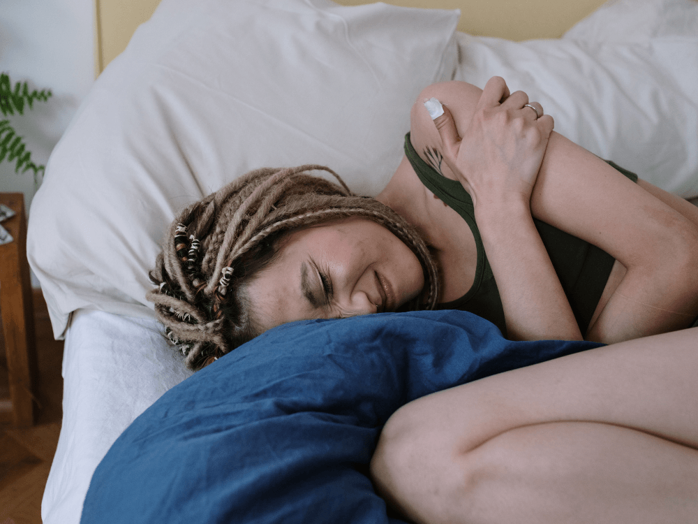 Mulher deitada de lado na cama, com os olhos fechados e uma mão sobre o abdômen, demonstrando possível desconforto abdominal.