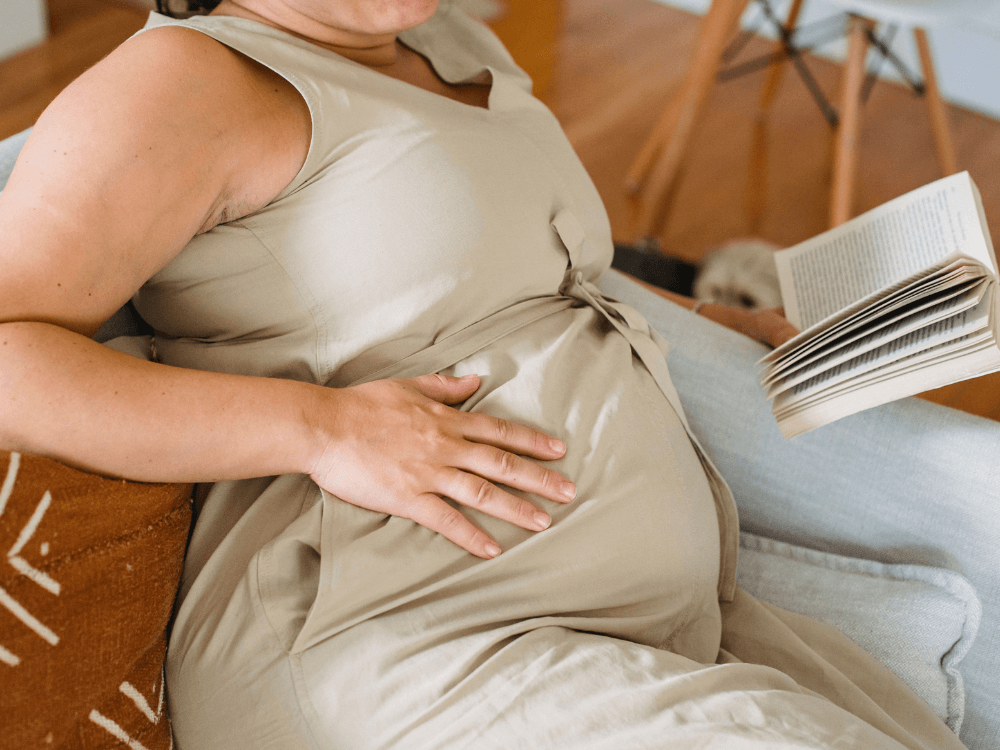 Mulher sentada no sofá mantém a mão sobre o abdômen volumoso, ilustrando o desconforto abdominal associado à retocolite ulcerativa. Tudo para explicar se retocolite ulcerativa incha a barriga.