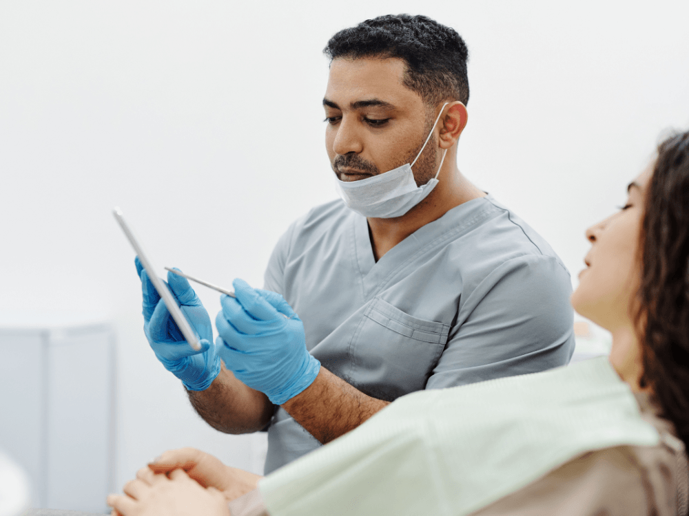 Médico com luvas azuis consulta informações em um dispositivo enquanto conversa com uma paciente durante atendimento clínico.